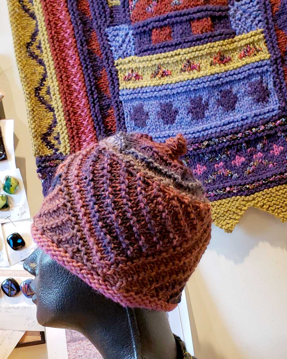 A knitted hat with a knitted wall hanging in the background. 